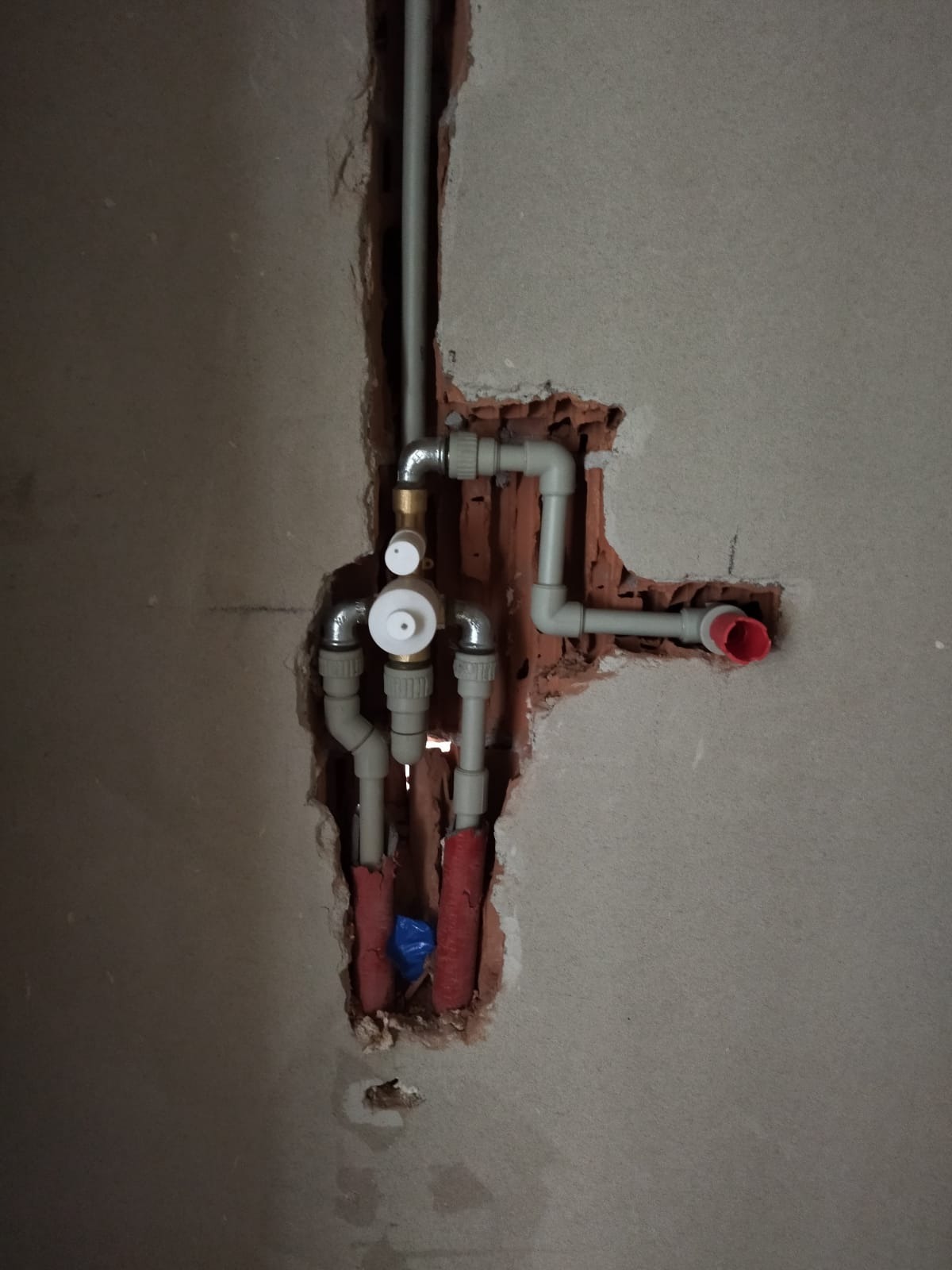 Fragment ściany z widoczną instalacją hydrauliczną w trakcie montażu, z rurami PEX owiniętymi czerwoną izolacją i elementami mocującymi, osadzonymi w wykuciu w ceglanej ścianie.