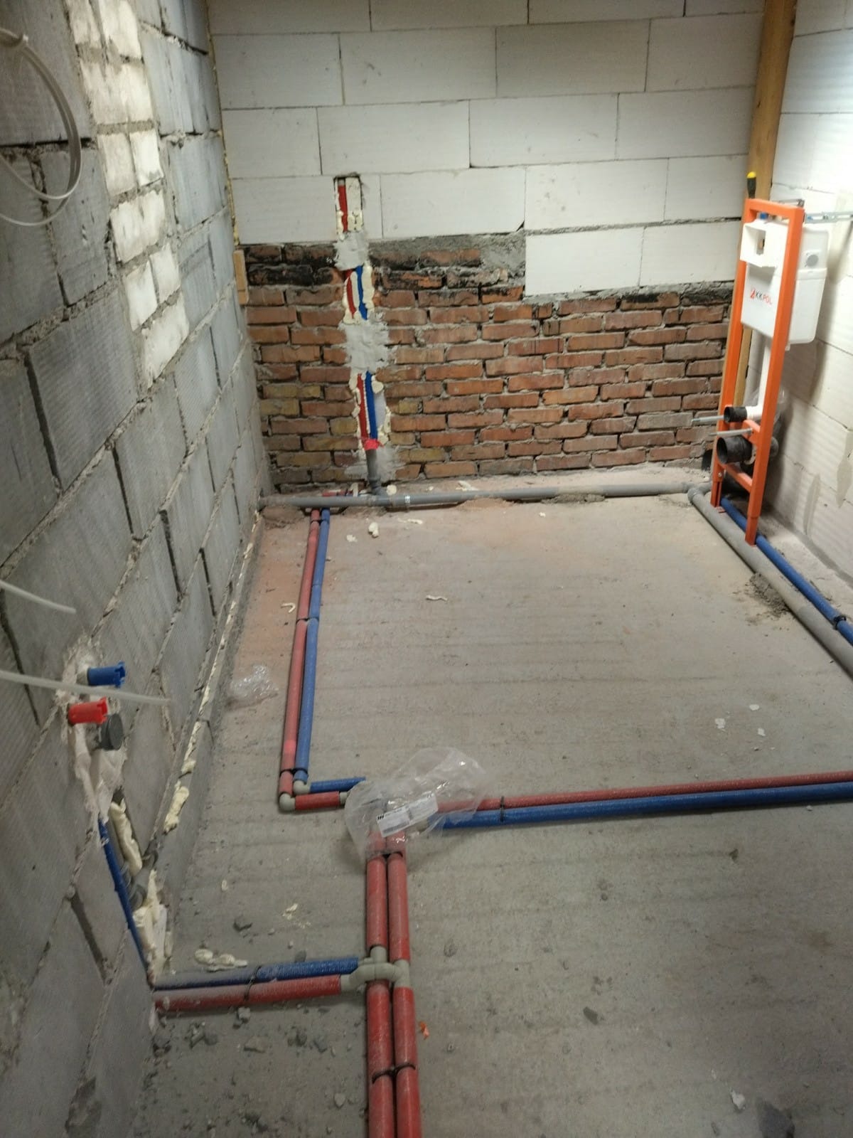 Surowy stan łazienki w trakcie prac hydraulicznych: widoczne czerwone i niebieskie rury prowadzone po podłodze i w ścianach z betonu komórkowego i cegły, zamocowany stelaż podtynkowy WC.