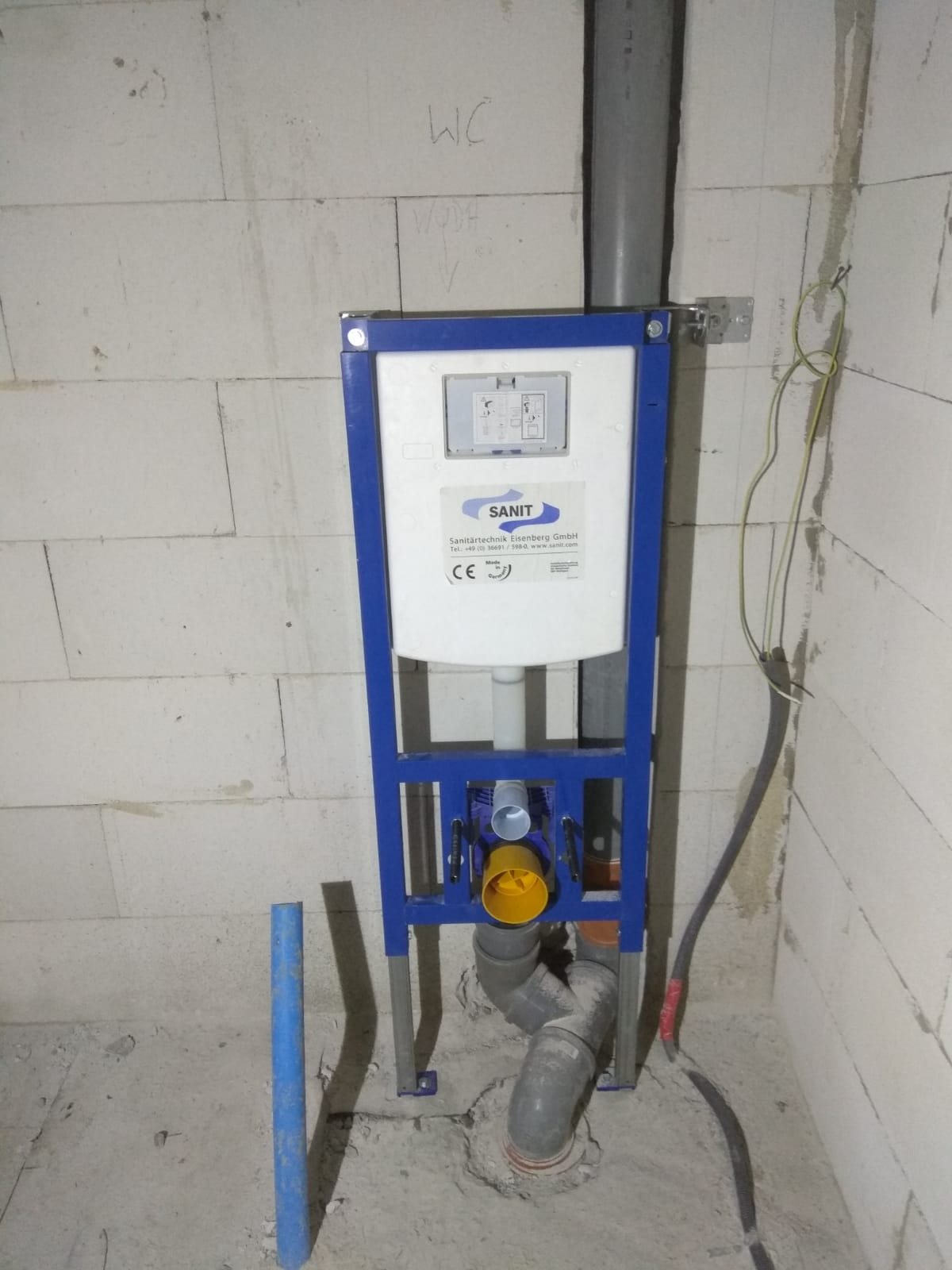 Stelaż podtynkowy WC marki Sanit z widocznymi przyłączami kanalizacyjnymi i rurą doprowadzającą wodę, zamontowany przy ścianie z betonu komórkowego z napisem 'WC'.