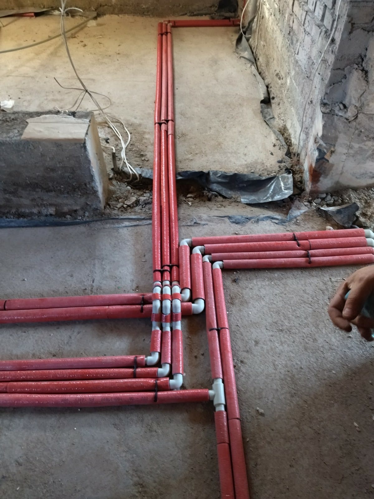 Rozłożone na surowym podłożu, czerwone rury w izolacji z białymi łącznikami tworzą skomplikowaną sieć instalacji podłogowej, zabezpieczone czarnymi opaskami zaciskowymi.