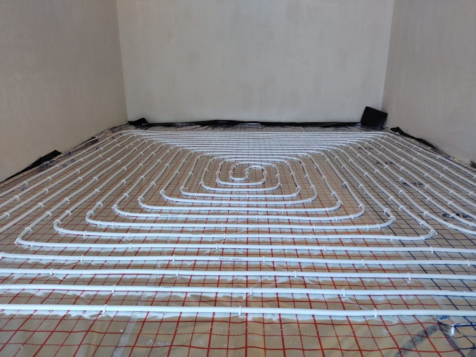 Białe rury ogrzewania podłogowego rozłożone spiralnie na folii z czerwoną kratką, widok z góry na surowy beton w tle.