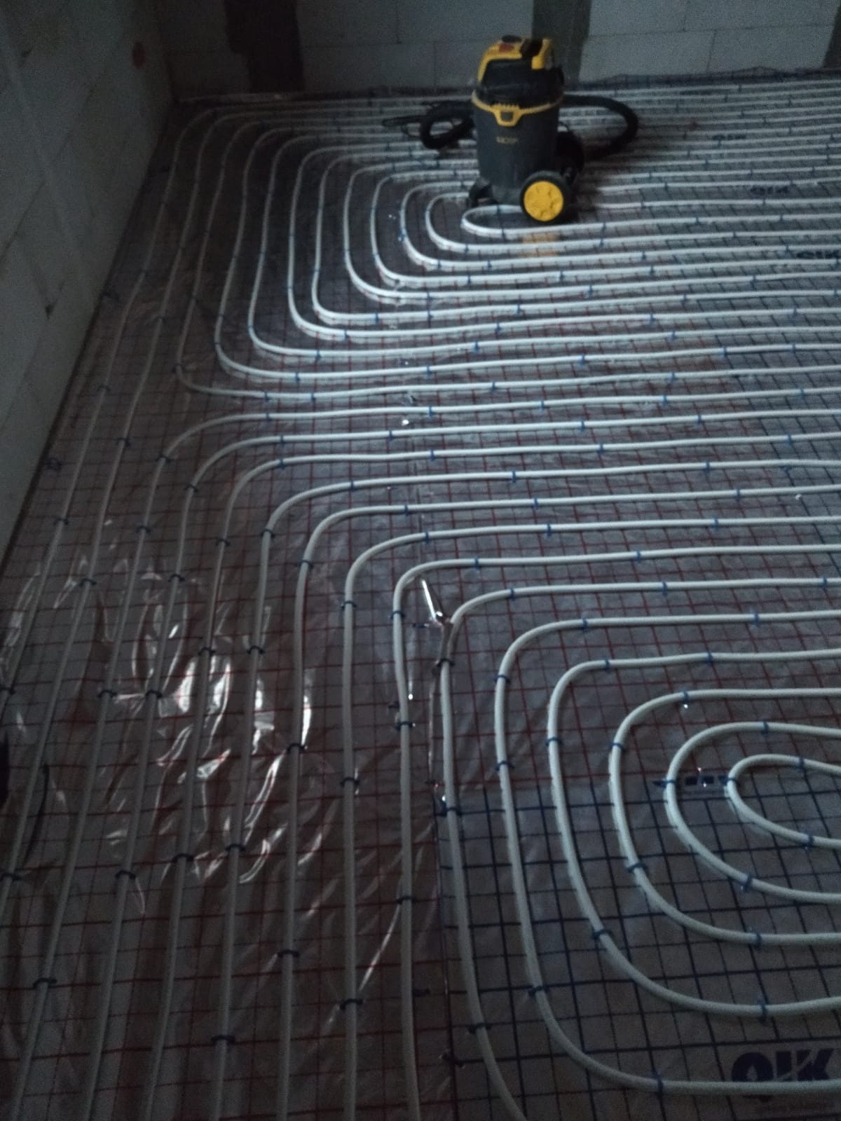 Białe rury ogrzewania podłogowego ułożone na siatce, widoczny odkurzacz przemysłowy w tle, surowy stan posadzki.