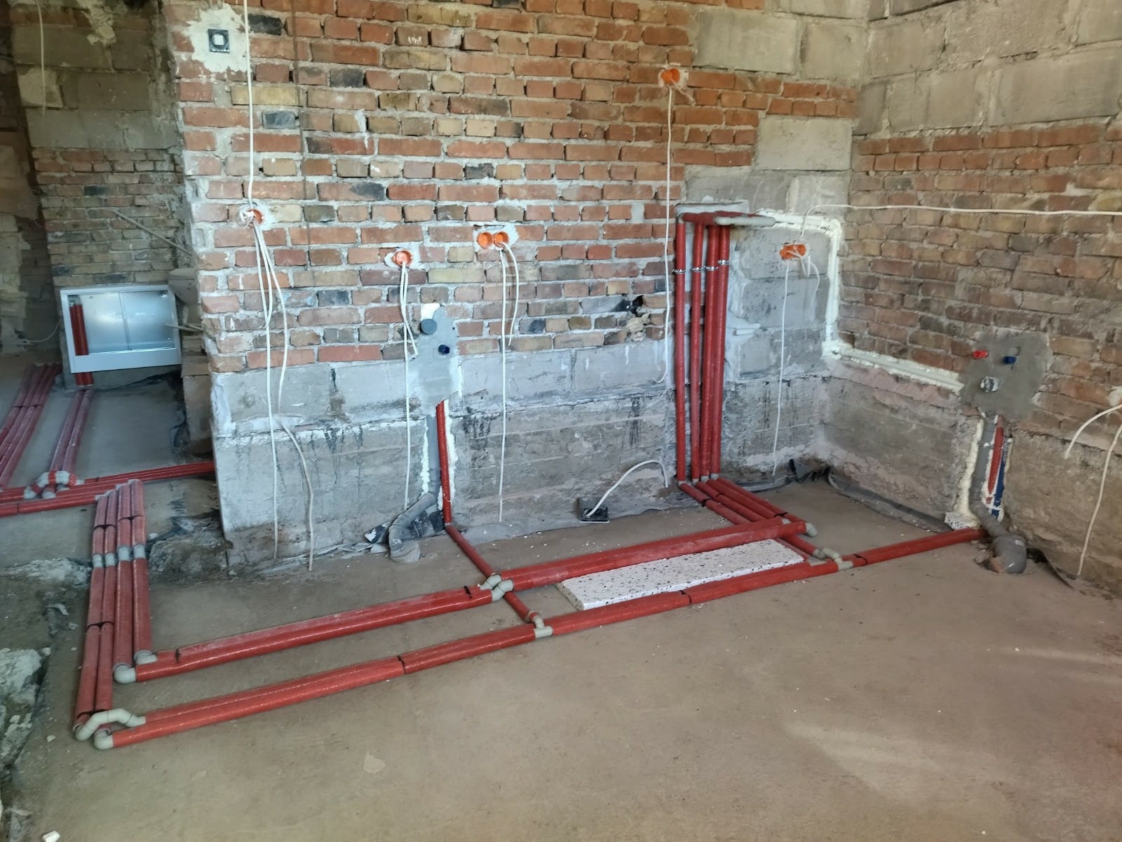 Czerwone rury instalacji wodnej ułożone na podłodze w surowym pomieszczeniu z ceglanymi ścianami i wystającymi przewodami elektrycznymi ze ściany.