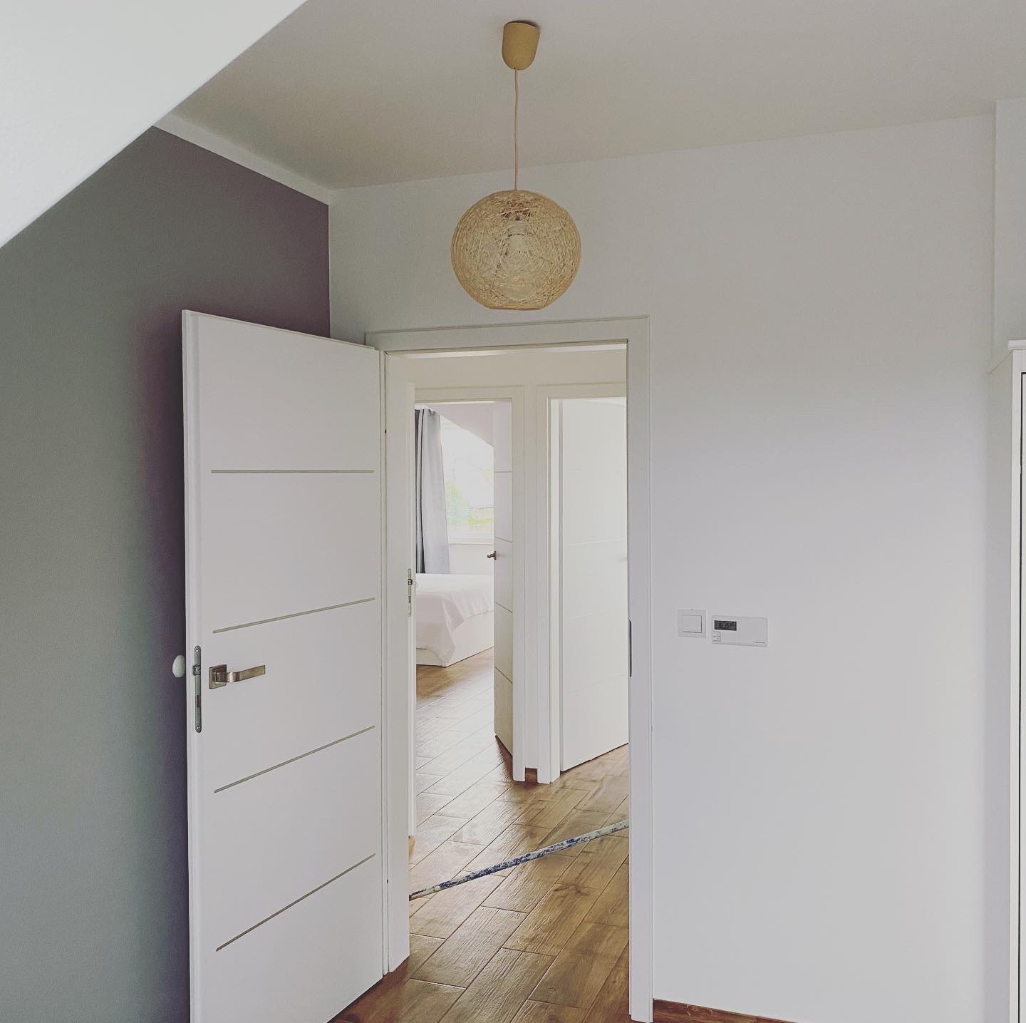 Wnętrze pokoju po remoncie: białe ściany, szara ściana akcentująca, drewniana podłoga, otwarte białe drzwi, widok na sypialnię z białym łóżkiem, pleciona lampa wisząca.