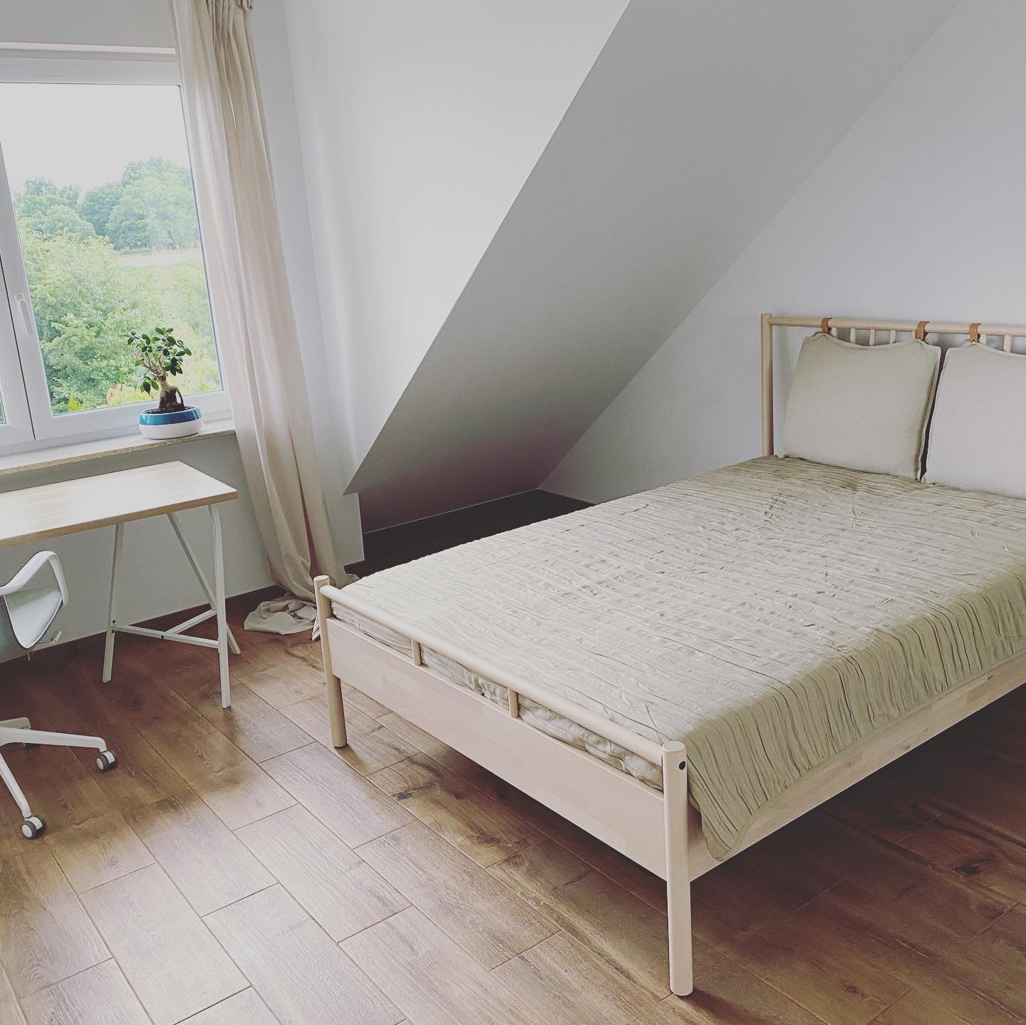 Jasne wnętrze pokoju z łóżkiem, biurkiem i oknem, drewniana podłoga, skośny sufit, minimalistyczny styl.