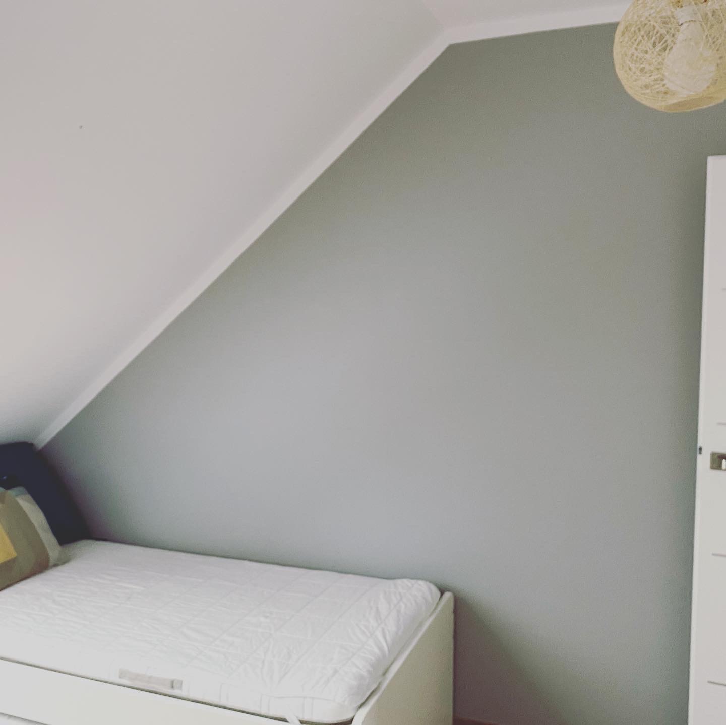 Pokój na poddaszu po remoncie, ściana pomalowana na szaro, biały sufit, białe łóżko z kołdrą w kratkę, lampa z abażurem z naturalnego sznurka.