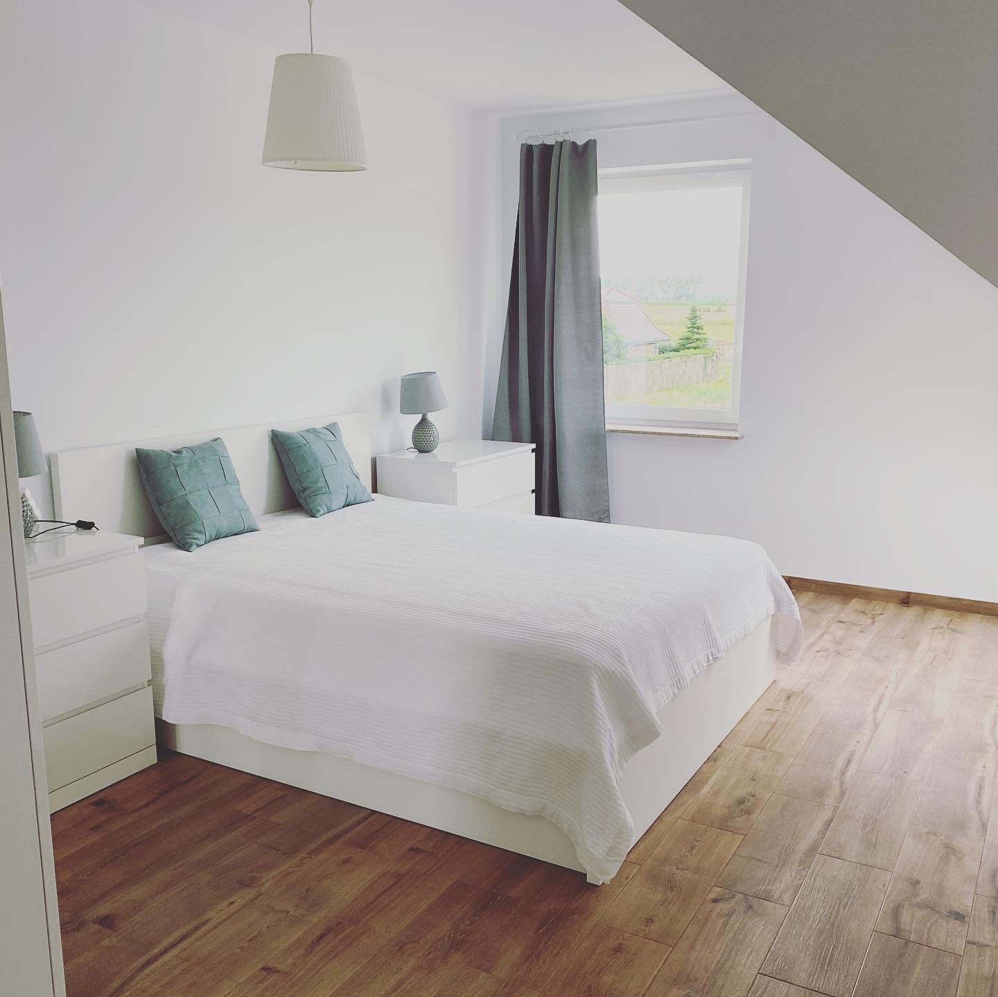 Jasna sypialnia na poddaszu z drewnianą podłogą, białymi ścianami, białym łóżkiem z turkusowymi poduszkami i szarą zasłoną w oknie.