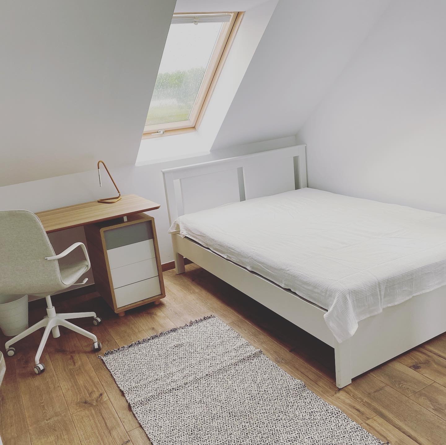 Jasne wnętrze pokoju na poddaszu z białymi ścianami, drewnianą podłogą, biurkiem, łóżkiem i oknem dachowym.