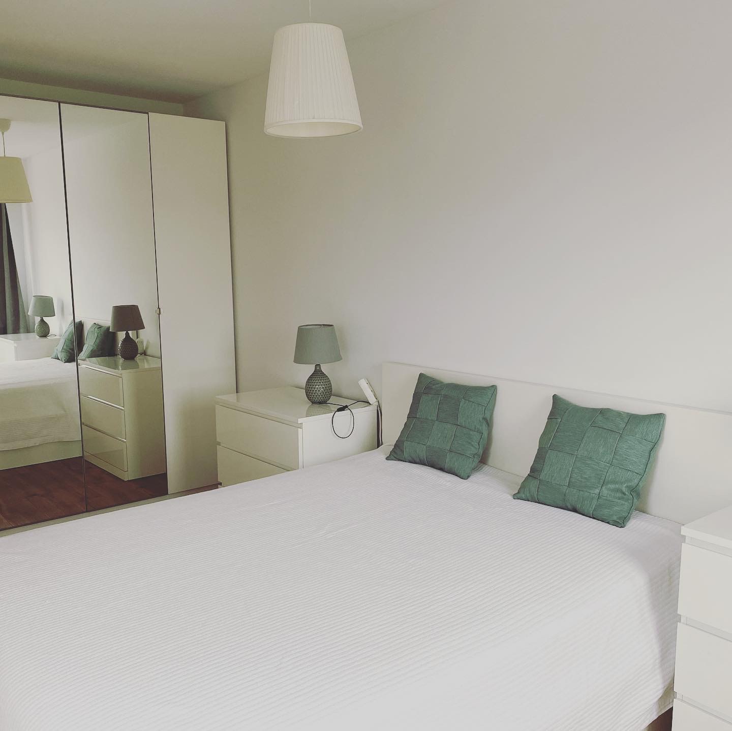Jasna sypialnia z białymi ścianami, łóżkiem z zielonymi poduszkami i szafą z lustrzanymi drzwiami, widoczna lampka nocna na szafce.