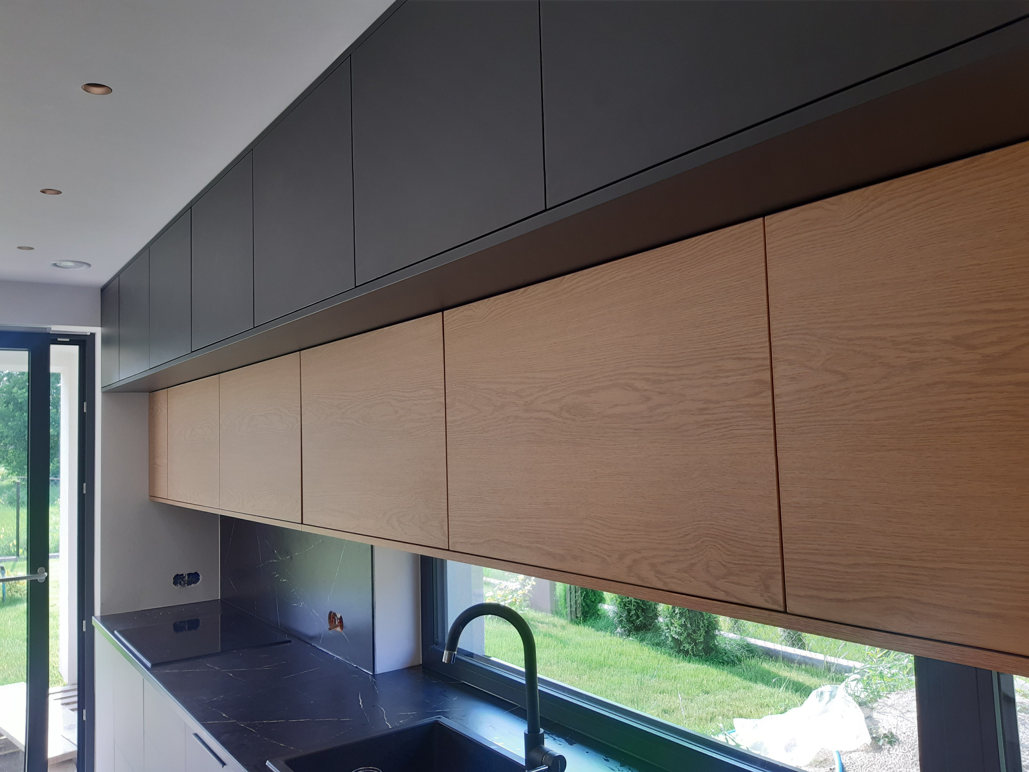 Nowoczesna kuchnia z górnymi szafkami w dwóch kolorach: grafitowym i drewnopodobnym, czarny blat z marmurkowym wzorem i widokiem na zieleń za oknem.