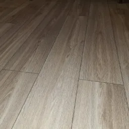 Jasne panele podłogowe z widoczną strukturą drewna, ułożone wzdłuż pomieszczenia, z widocznymi łączeniami i minimalnym oświetleniem.