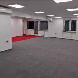 Wnętrze biura po remoncie: szara wykładzina dywanowa, białe ściany, klimatyzatory i kwadratowe panele LED w suficie, widoczne okna z grzejnikami.