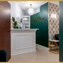 Wnętrze kliniki medycyny regeneracyjnej: biała recepcja z rośliną doniczkową, ściana w kolorze butelkowej zieleni z logo, kryształowy żyrandol, wzorzysta tapeta, ciemne drzwi wejściowe i fragment...