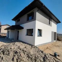 Dwukondygnacyjny dom jednorodzinny z szarą górną i białą dolną częścią elewacji, czarnym dachem i oknami, otoczony ziemią i piaskiem, błękitne niebo w tle.