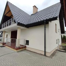 Dis-Bud Damian Żmij - Wykończona elewacja domu jednorodzinnego z balkonem, widoczne orynnowanie i fragment kostki brukowej, pochmurne niebo.