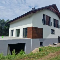 Dis-Bud Damian Żmij - Dwukondygnacyjny dom z białymi ścianami i szarą podmurówką, z drewnianymi akcentami na elewacji i oknach, widoczny z perspektywy trawnika przed budynkiem.