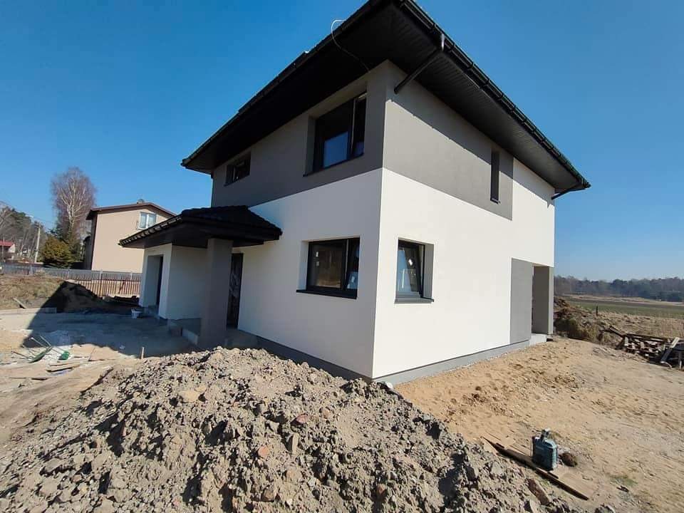 Dwukondygnacyjny dom jednorodzinny z szarą górną i białą dolną częścią elewacji, czarnym dachem i oknami, otoczony ziemią i piaskiem, błękitne niebo w tle.