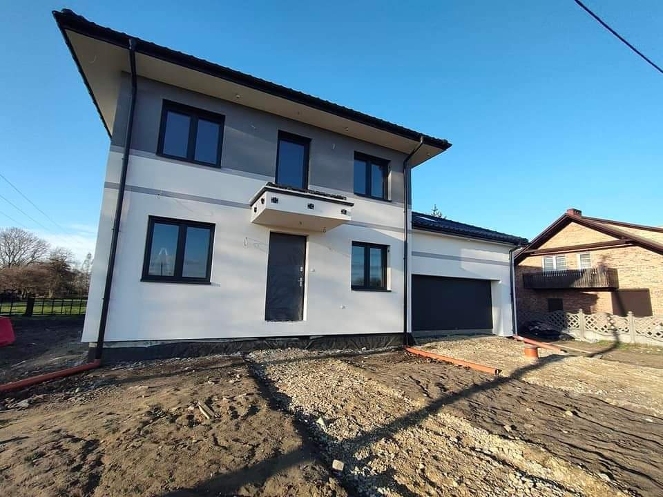 Dwukondygnacyjny dom z elewacją w odcieniach bieli i szarości, czarne okna i drzwi, teren wokół domu w trakcie zagospodarowania, widoczne rury odprowadzające wodę.