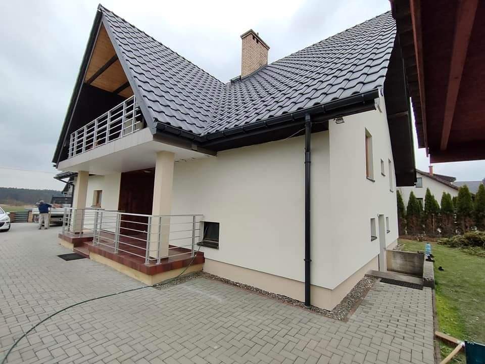 Wykończona elewacja domu jednorodzinnego z balkonem, widoczne orynnowanie i fragment kostki brukowej, pochmurne niebo.