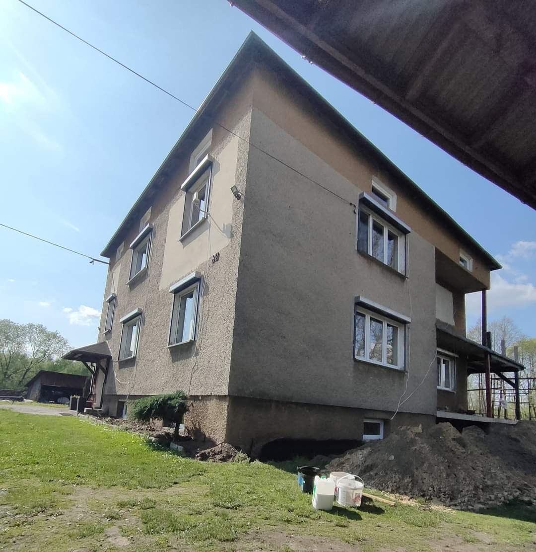 Dwukondygnacyjny dom z szarą elewacją, częściowo pomalowany na jasny kolor, widoczne okna z roletami, sterty ziemi i puste wiadra po farbie na trawniku.