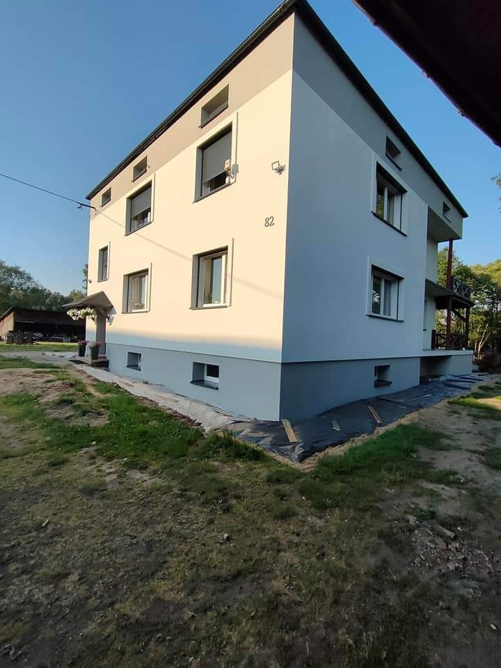 Dwukondygnacyjny budynek z numerem 82, świeżo pomalowaną elewacją w odcieniach szarości i bieli, widoczny z niskiej perspektywy, z fragmentem rozłożonej folii ochronnej u podstawy.