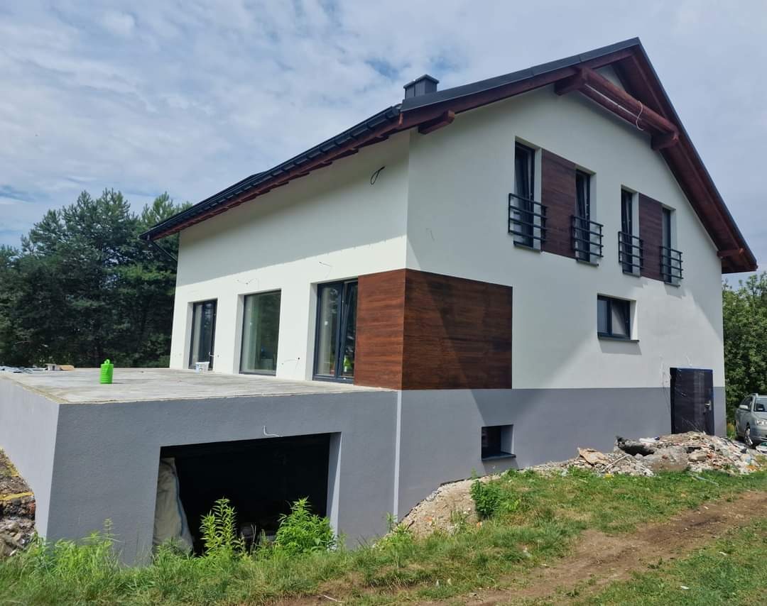 Dwukondygnacyjny dom z białymi ścianami i szarą podmurówką, z drewnianymi akcentami na elewacji i oknach, widoczny z perspektywy trawnika przed budynkiem.