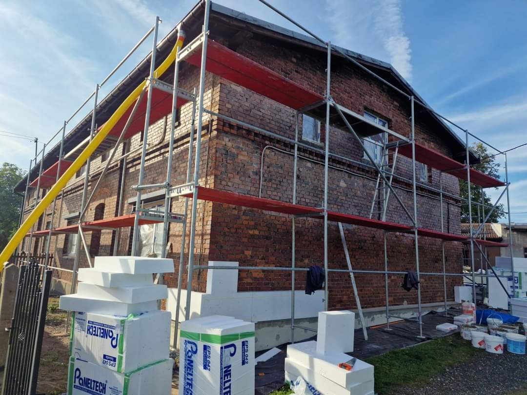 Ceglany budynek w trakcie prac, widoczne rusztowania, częściowo przyklejony styropian izolacyjny, paczki styropianu i wiaderka z klejem na trawniku.