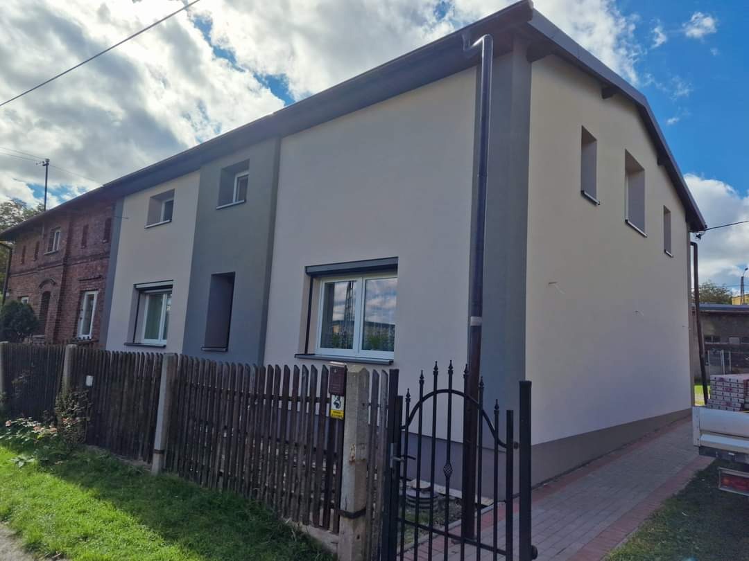 Dwukondygnacyjny budynek po renowacji elewacji, z nową, jasną kolorystyką i odnowioną stolarką okienną, widoczny zza drewnianego płotu i metalowej bramki.