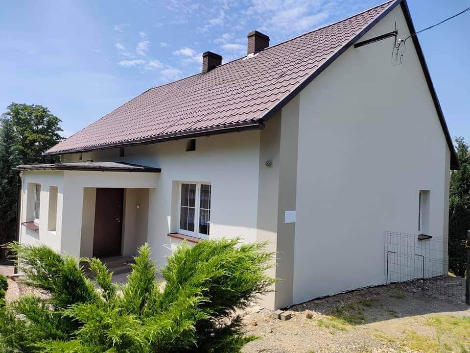 Dom jednorodzinny z nową, jasną elewacją i brązowym dachem, widoczny z perspektywy ogrodu z krzewami iglastymi na pierwszym planie.