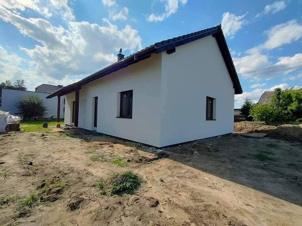 Świeżo pomalowany, parterowy dom z ciemną dachówką i brązowymi oknami, widoczny z boku na tle błękitnego nieba z chmurami. Teren wokół domu jeszcze niezagospodarowany, widoczne ślady prac ziemnych.