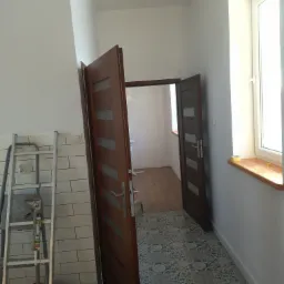 Wnętrze po remoncie: białe ściany, wzorzysta podłoga, drewniane drzwi z przeszkleniami, drabina oparta o ścianę z białymi płytkami.