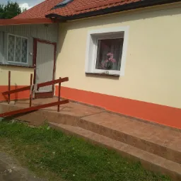 Świeżo pomalowana fasada domu z oknem, w którym widać storczyki, z pomalowanym na pomarańczowo cokołem i schodami wyłożonymi płytkami imitującymi marmur.