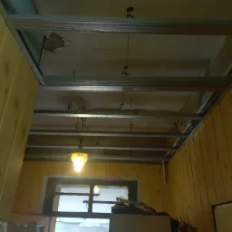 Montaż metalowej konstrukcji sufitu podwieszanego w pomieszczeniu z boazerią, widoczna stara instalacja elektryczna i wisząca lampa.