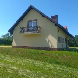 Jasna elewacja domu jednorodzinnego z balkonem i ceglanym cokołem, widok z boku na tle błękitnego nieba i skoszonej trawy.