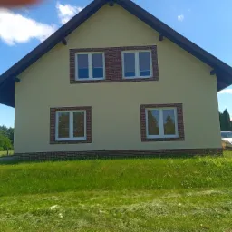 Dwukondygnacyjny dom z ocieploną elewacją w odcieniach beżu, z brązowymi obramowaniami okien i dachu, otoczony zielonym trawnikiem i błękitnym niebem z widocznymi chmurami, z zaparkowanym...