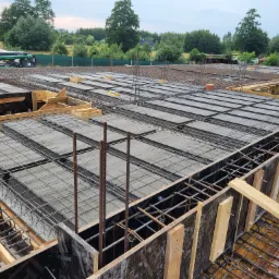Zbrojenie stropu budynku w trakcie betonowania, widoczne szalunki, stalowe pręty i siatka zbrojeniowa, w tle zieleń drzew.