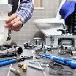 Instalator hydraulik montuje biały syfon umywalkowy, widoczne narzędzia, chromowane elementy instalacji, płytki ceramiczne i toaleta w tle.