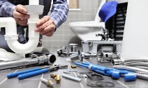 Instalator hydraulik montuje biały syfon umywalkowy, widoczne narzędzia, chromowane elementy instalacji, płytki ceramiczne i toaleta w tle.