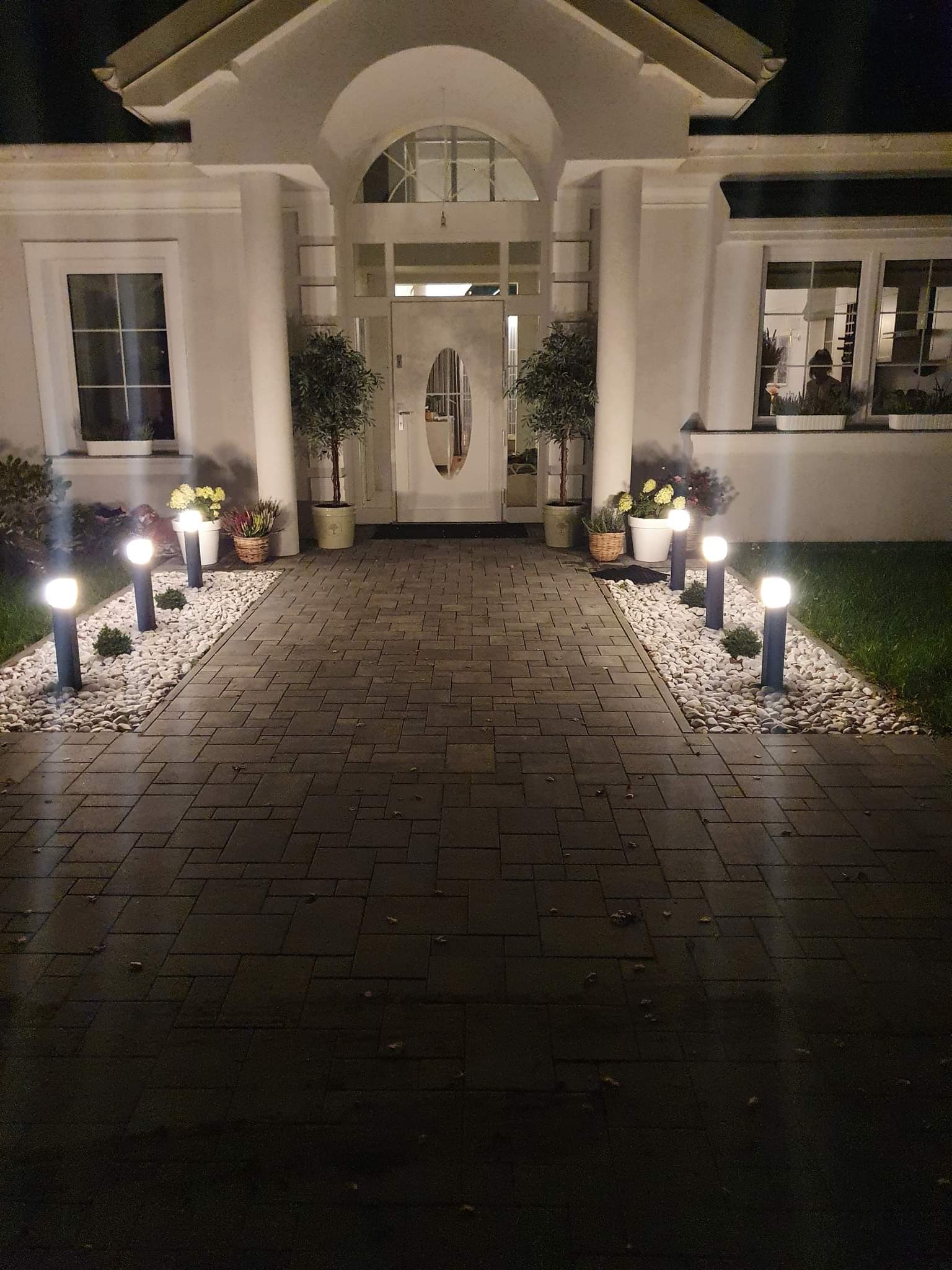 Oświetlona ścieżka z kostki brukowej prowadząca do wejścia do domu z kolumnami, oświetlona lampami ogrodowymi i wysypana białym kamieniem.