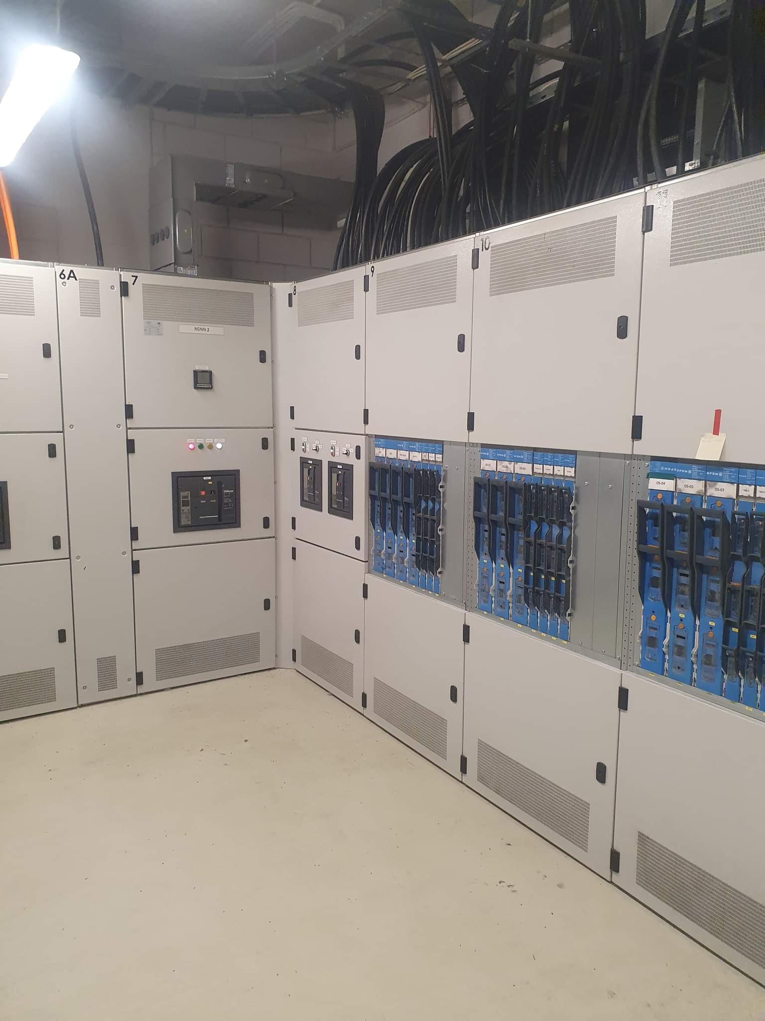 Rozbudowana rozdzielnica elektryczna z modułami w kolorze niebieskim, umieszczona w pomieszczeniu technicznym z widocznymi kablami na suficie.