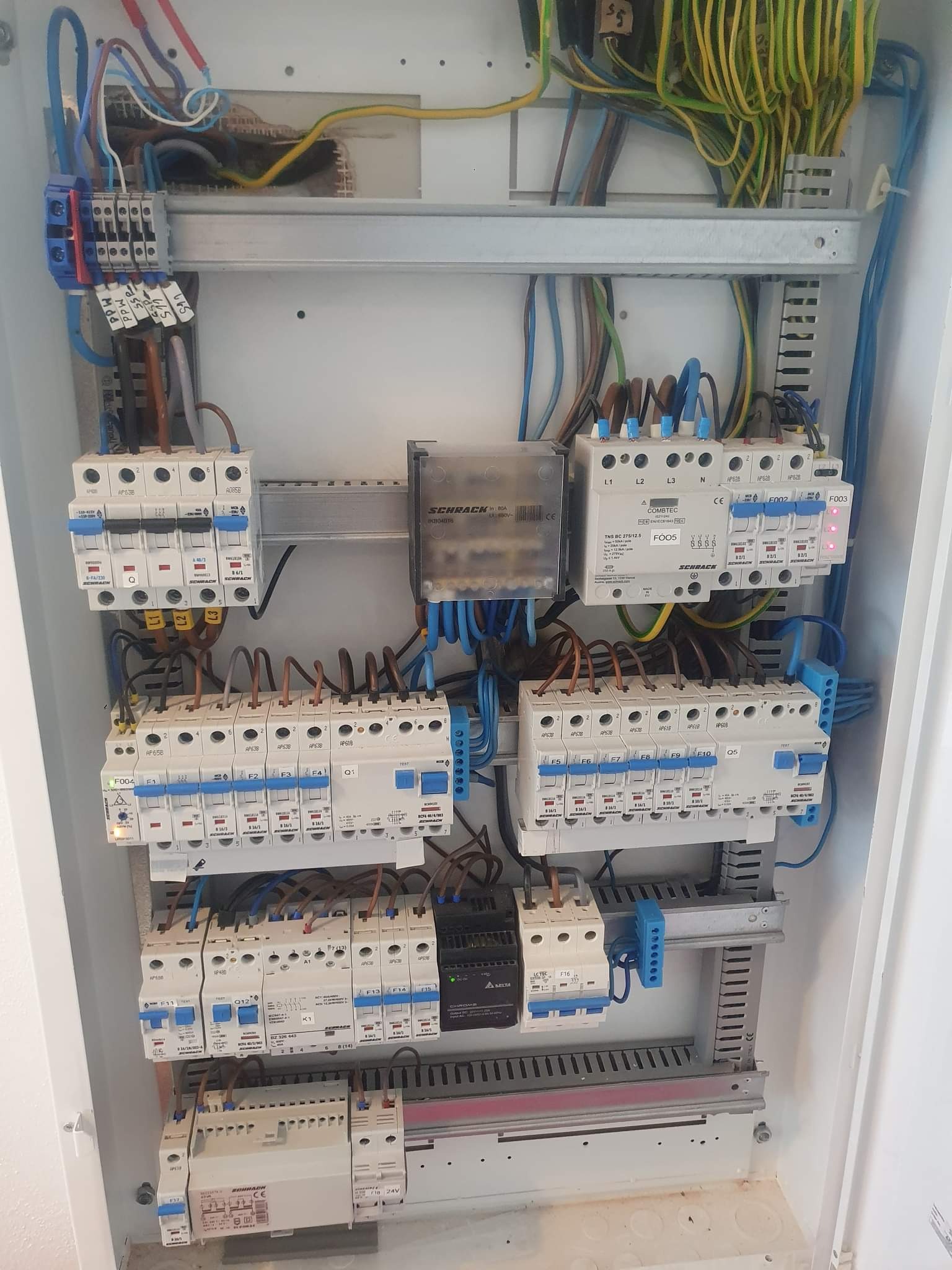 Otwarta skrzynka elektryczna z zamontowanymi bezpiecznikami, różnymi przewodami i elementami elektrycznymi. Widoczne oznaczenia na bezpiecznikach i przewodach. Górna część skrzynki z licznymi kablami.