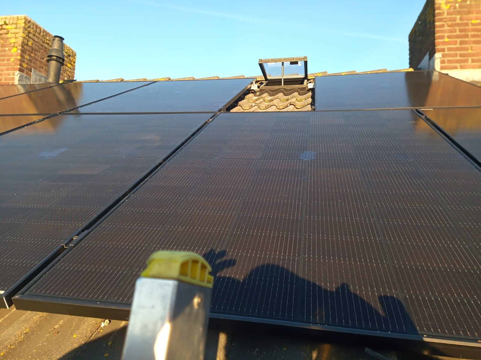 Panele fotowoltaiczne na dachu domu z ceglanym kominem w tle, widoczne z perspektywy drabiny z żółtym zakończeniem, z otwartym oknem dachowym.