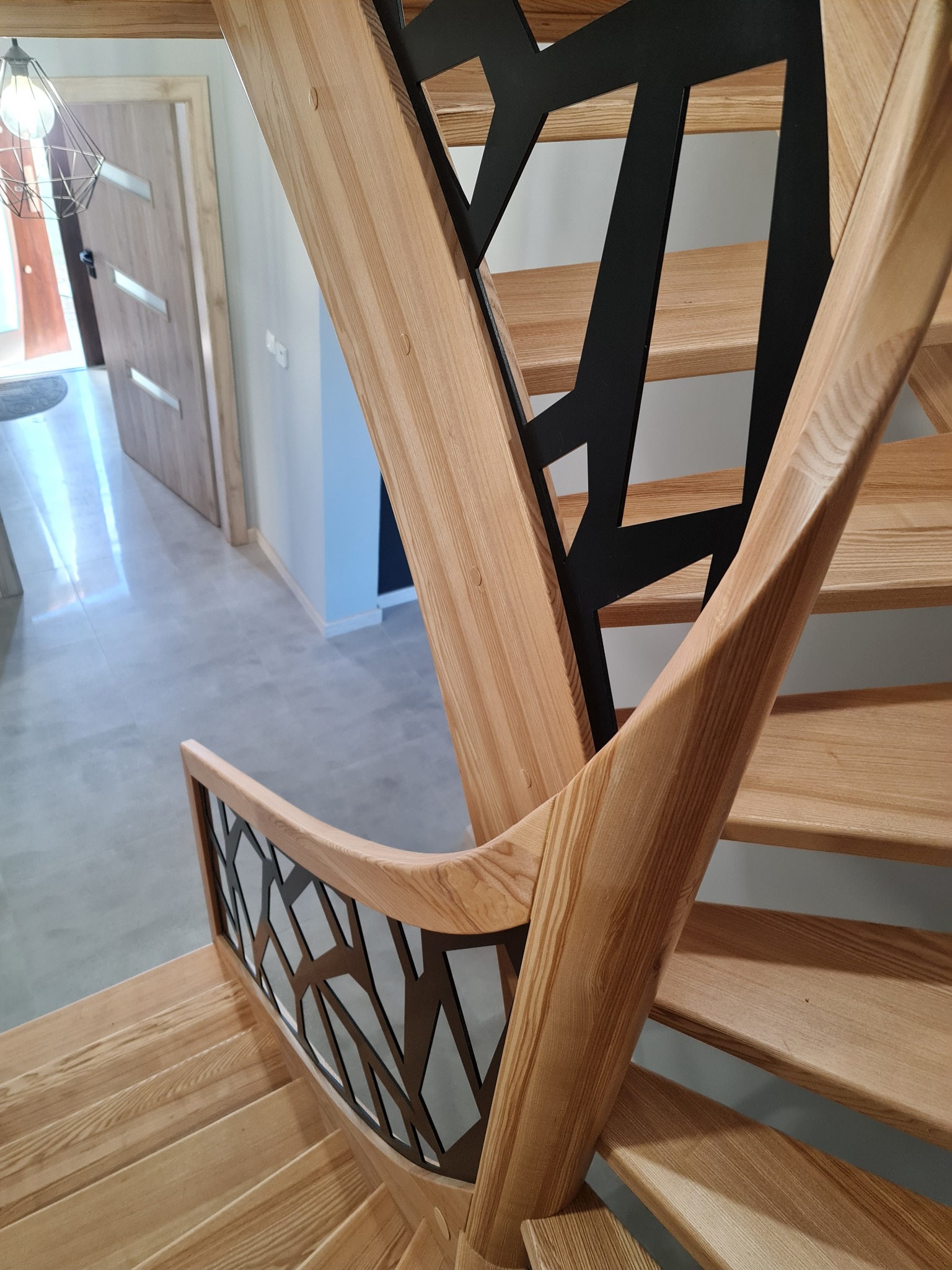Drewniane schody z jasnego drewna z czarną, geometryczną balustradą, widok z góry, w tle widoczna podłoga z płytek i drzwi.