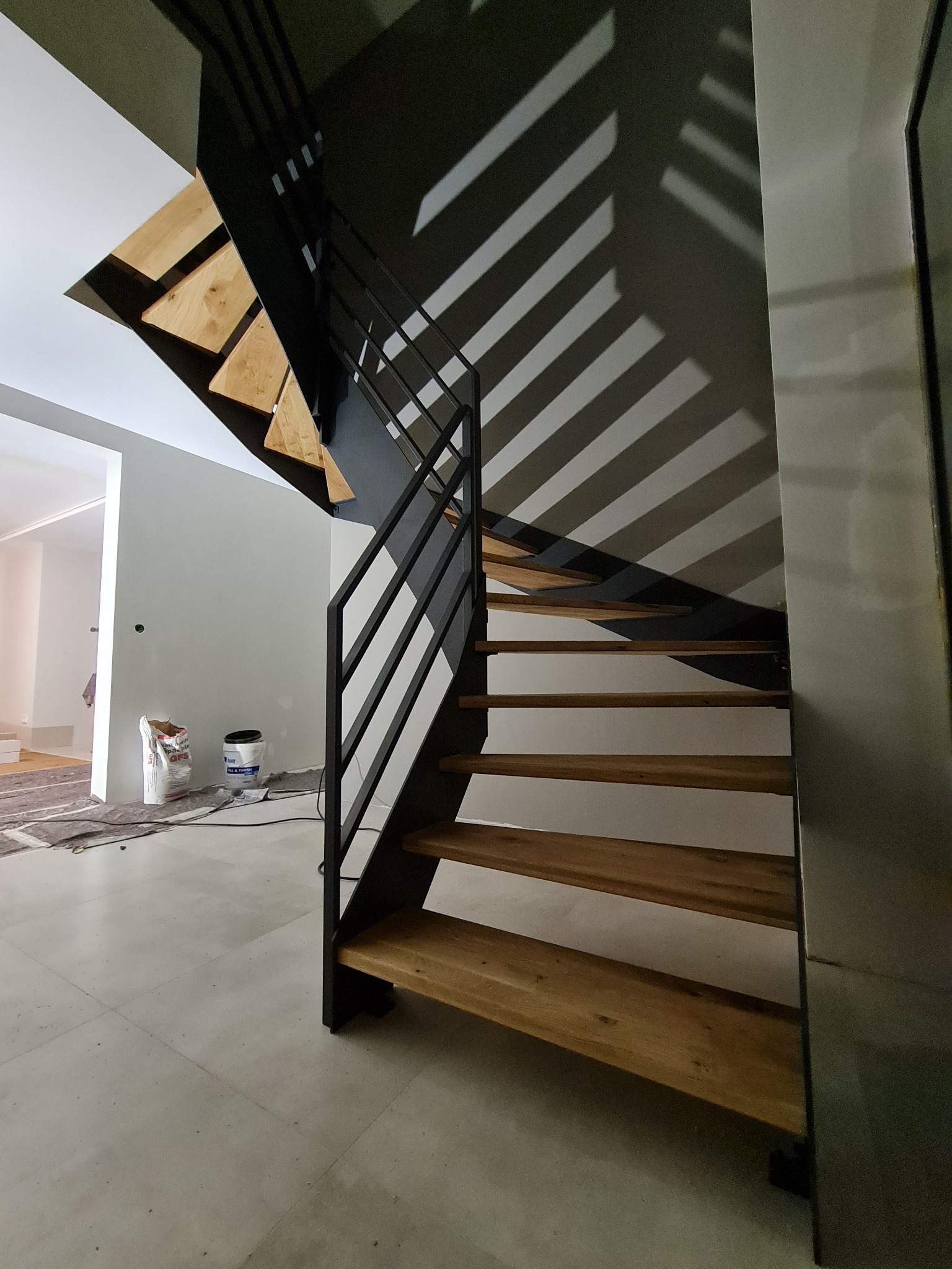 Nowoczesne, spiralne schody z drewnianymi stopniami i czarną, metalową konstrukcją, rzucające cienie na szarą ścianę. W tle widoczne materiały budowlane.