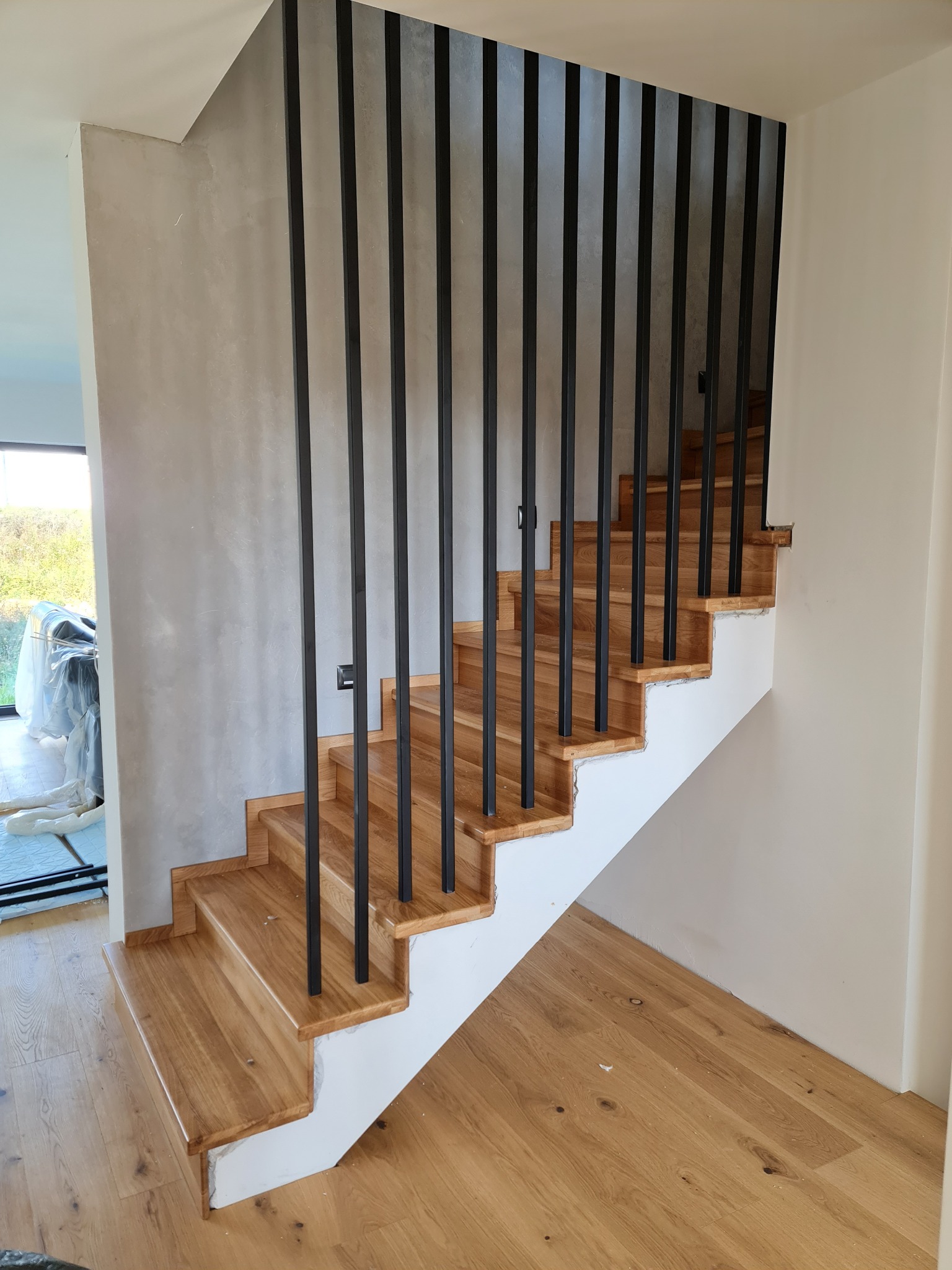 Nowoczesne schody z drewnianymi stopniami i czarnymi, pionowymi balustradami, widoczne niedokończone wykończenie ściany i podłogi.