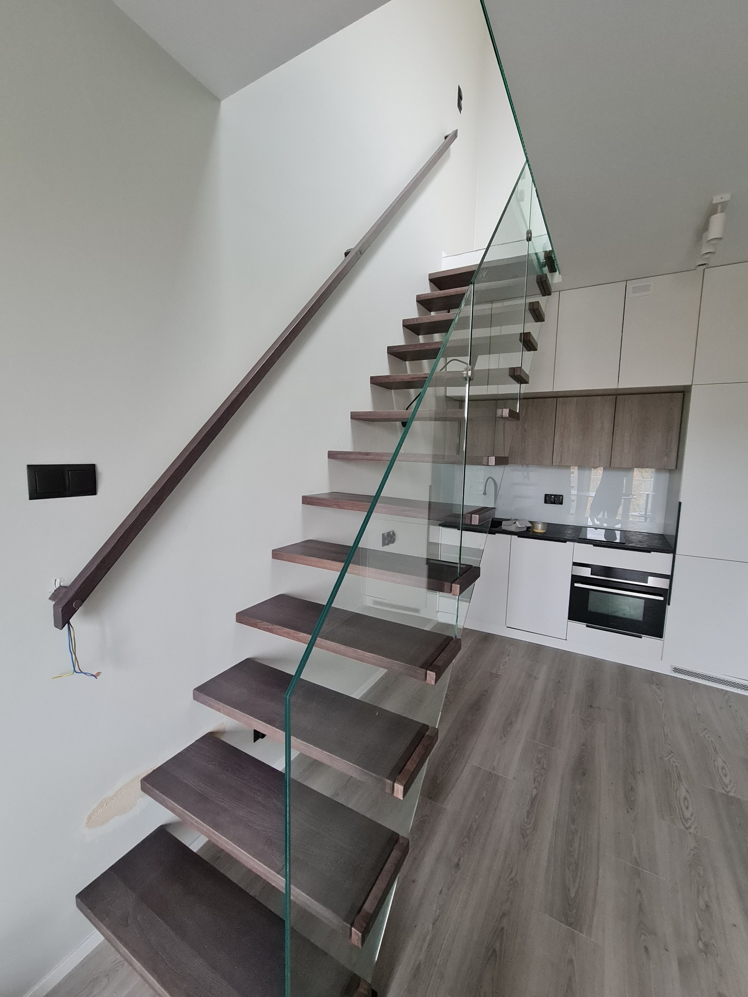 Nowoczesne schody z ciemnego drewna z szklaną balustradą, widok z dołu, w tle minimalistyczna kuchnia z białymi szafkami, widoczne niedokończone prace wykończeniowe przy ścianie.