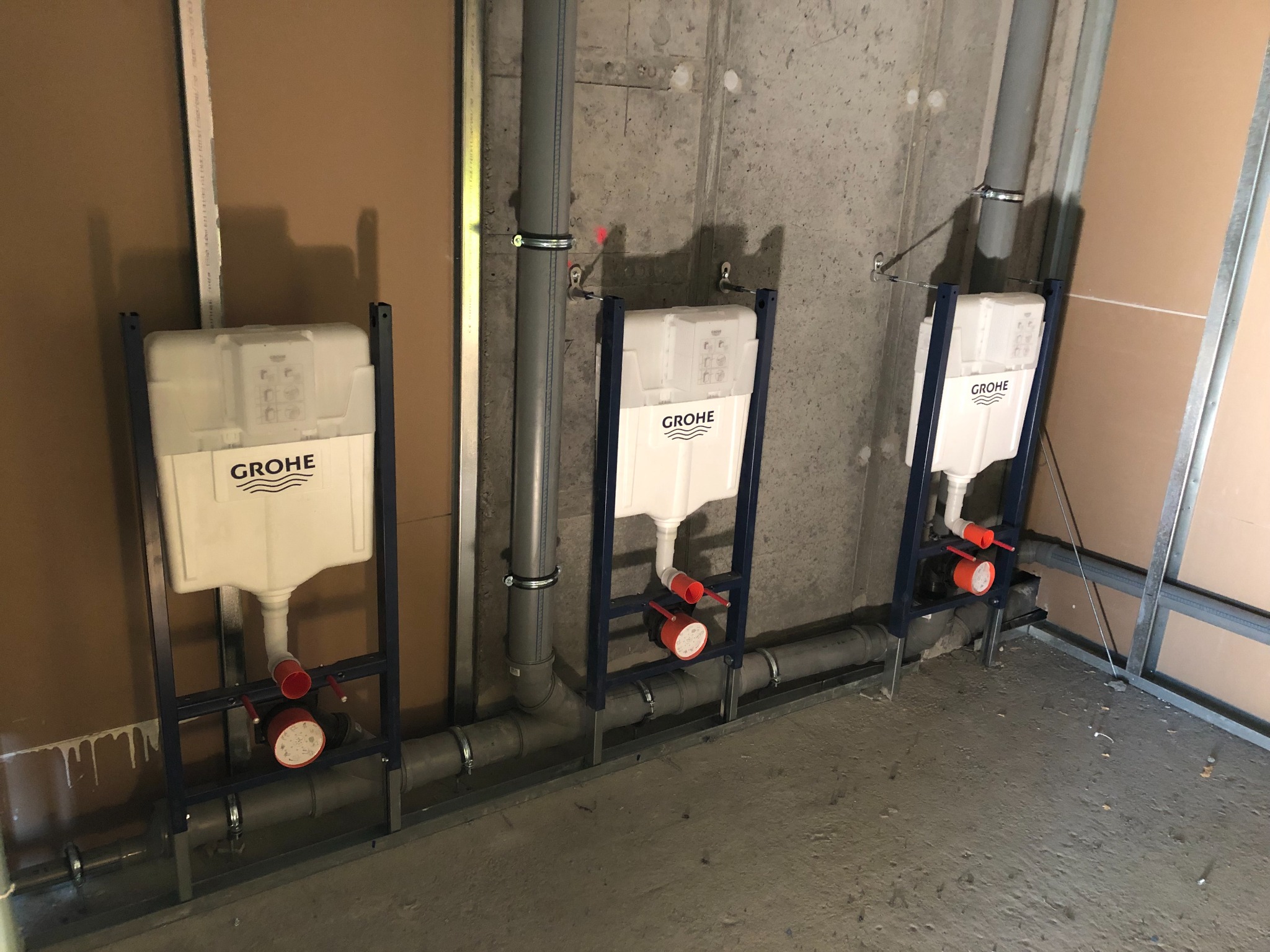 Trzy podtynkowe stelaże WC marki Grohe zamontowane na szarym betonowym murze, z widocznymi rurami kanalizacyjnymi i elementami instalacji wodnej, w surowym stanie wykończenia łazienki.