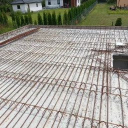 Zbrojenie stropu żelbetowego z widocznymi prętami stalowymi i betonem, przygotowane do zalania, z domami jednorodzinnymi w tle.