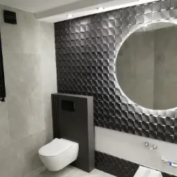 Nowoczesna łazienka z geometryczną ścianą z płytek 3D, podświetlanym okrągłym lustrem i wiszącą toaletą w zabudowie.