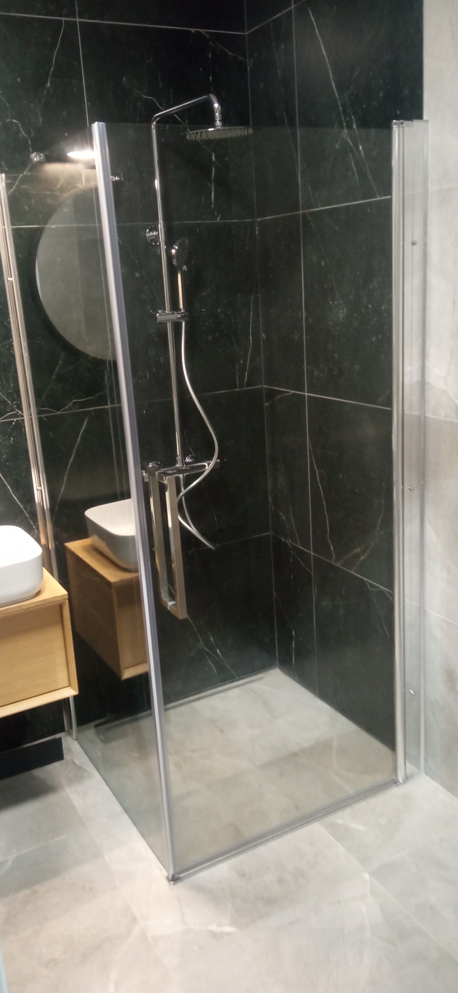 Nowoczesna kabina prysznicowa ze szklanymi drzwiami, czarne marmurowe płytki na ścianach, okrągłe lustro i drewniana szafka pod umywalką.